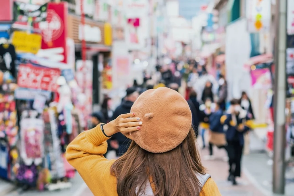 Woman in a beret and orange sweater facing away and looking over a bustling Tokyo shopping street, representing Japan market entry for fashion brands.